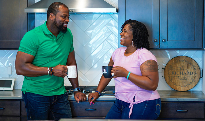 Warrior Taniki Richard stands in her kitchen with her husband while drinking from coffee mugs and smiling at each other.