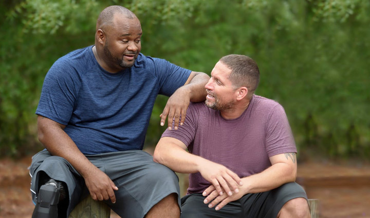 Wounded warriors Chris Gordon and Manny Colon talk with each other while sitting on tree stumps outside.
