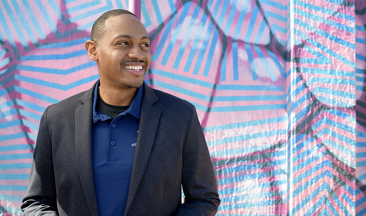 Wounded warrior Tyshawn Jenkins stands in front of an abstract pink and blue mural while looking to his left and smiling at someone out of frame. 