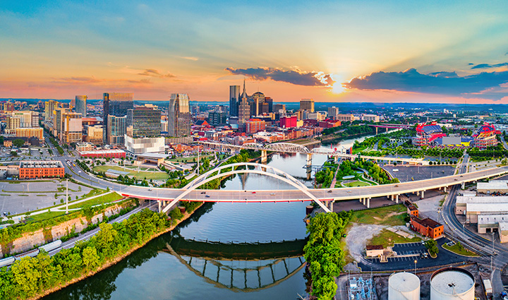 Aerial sunset view of Nashville with the AT&T Building, pedestrian bridge, river reflections, and warm sky tones.