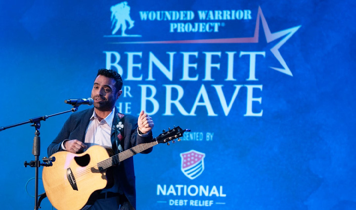 Wounded warrior Sal Gonzalez performs in a suit with a guitar around his neck against a blue backdrop that says “Benefit for the Brave”.