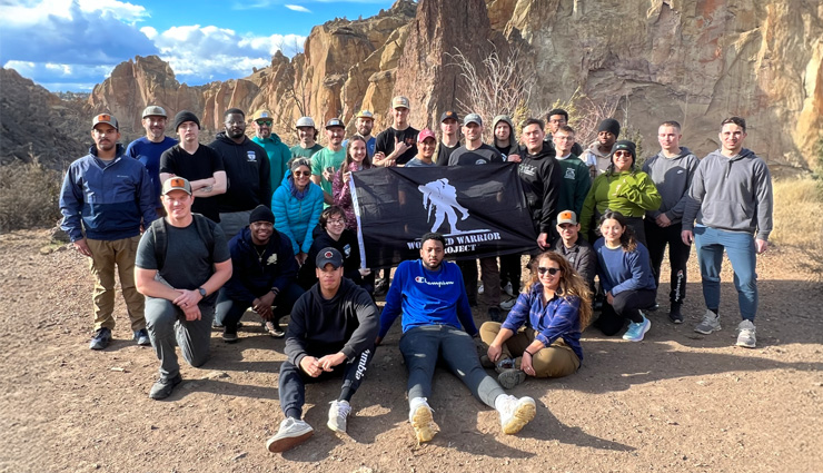 A group of wounded warriors gather for a group photo in front of some mountains while holding a black flag with a white WWP logo. 