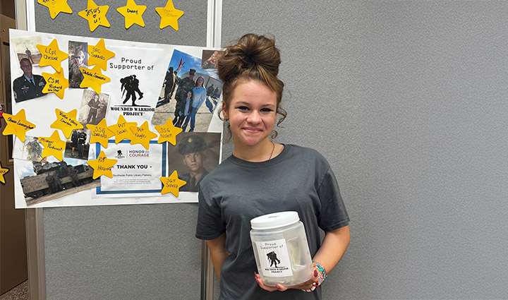 An Honor Their Courage student holds a donation jar for WWP. She stands next to a tribute board of photos and names of veterans.