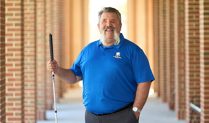 Wounded warrior Aaron Cornelius stands in the hallway of a brick building while smiling with a walking stick in hand.