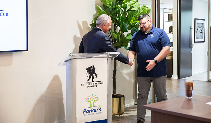 Greg Parker, Founder and CEO of Parker’s Kitchen, is wearing a gray jacket while shaking hands with a wounded warrior Jake Norotsky as he comes to the podium.
