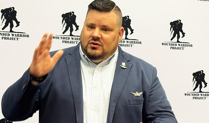 Veteran Jake Norotsky speaks on stage in a blue suit jacket in front of a white backdrop with WWP logos.