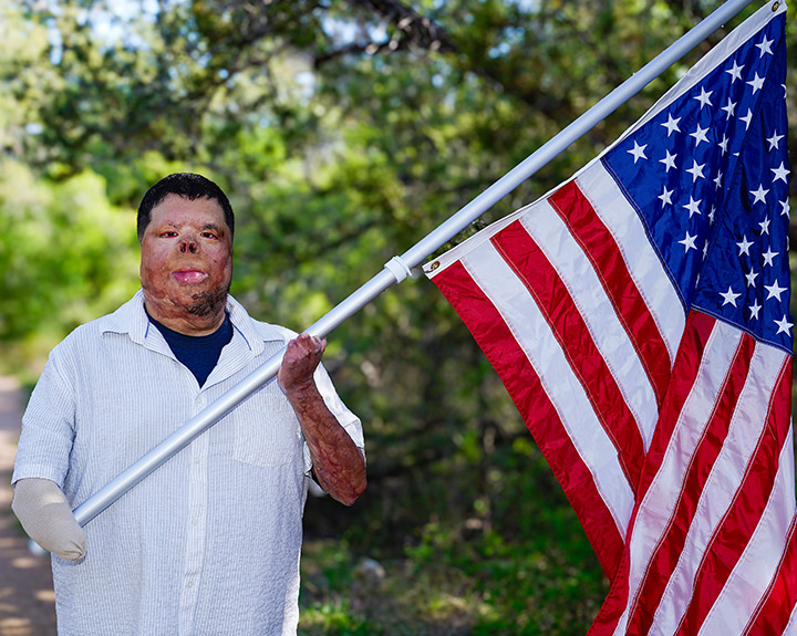 Wounded warrior Anthony Villareal stands on a tree-lined path while wearing a white button-down shirt and holding an American flag.