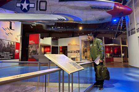 Warrior Tyshawn Jenkins, in a flight jacket, reads a Tuskegee Airmen exhibit beneath a suspended WWII fighter plane.
