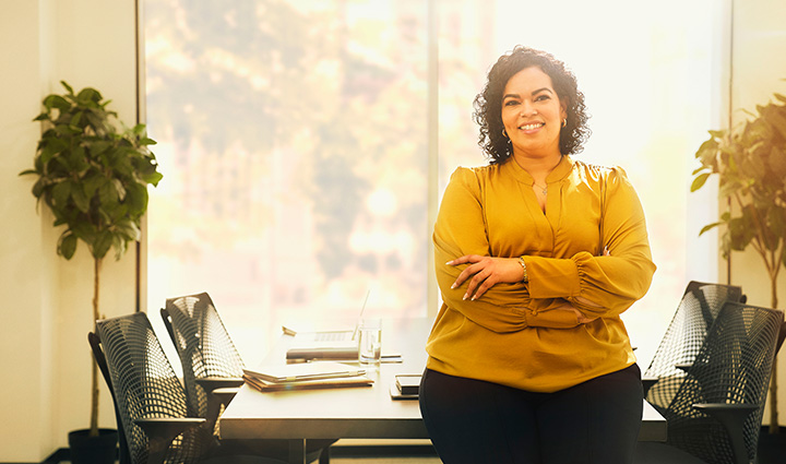 Wounded warrior Yomari Cruz stands with her arms crossed in a sunlit office, in front of a desk with papers, laptop, and plants. 