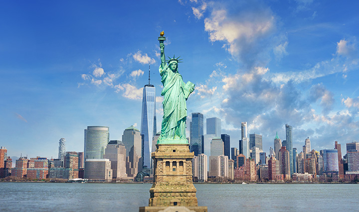 Statue of Liberty with NYC skyline in background, including One World Trade Center, under a clear sky across the water.