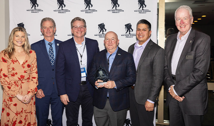 WWP leadership standing with USAA holding an award, smiling, wearing business attire standing in front of a WWP backdrop.