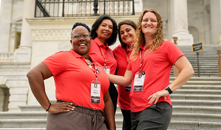 Four women warriors in matching WWP polo shirts stand on the steps of the U.S. Capitol building while they huddle together and smile at the camera.