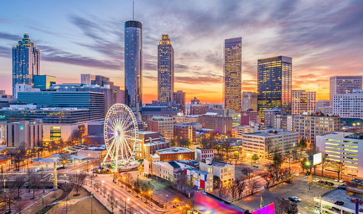 Atlanta skyline at sunset with tall buildings, Ferris wheel, and a sky glowing orange, pink, and purple above city lights. 